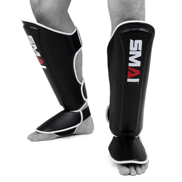 A person stands barefoot against a white background wearing black SMAI Essentials Muay Thai Shin Guards - Kids, featuring white trim and designed to cover the shins and instep for martial arts.