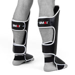 Two lower legs wearing black and white SMAI Essentials Muay Thai Shin Guards - Kids, featuring foot protection, standing on a white background.