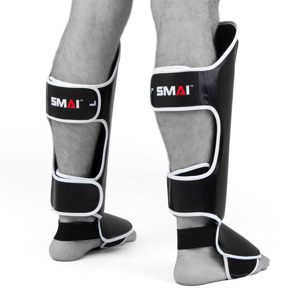Two lower legs wearing black and white SMAI Essentials Muay Thai Shin Guards - Kids, featuring foot protection, standing on a white background.