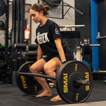 In a gym, a woman in black t-shirt and shorts prepares to deadlift a barbell loaded with SMAI HD Bumper Plates (Pair) - 15kg. Barefoot and focused, she highlights this essential strength training gear amid the equipment.