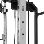 Close-up of a metal SMAI All-In-One Home Gym Trainer - Plate Loaded - Demo Stock frame, showing numbered adjustment holes (4, 5, 7). Cables and structural bars from this home gym are visible in the background.