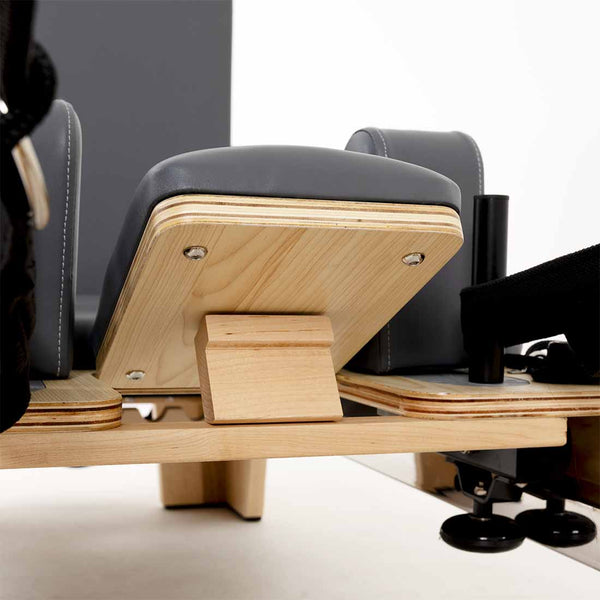 Close-up of the SMAI Pilates - Classical Maple Reformer, featuring a wooden base, padded gray cushions, metal bolts, and adjustable components.