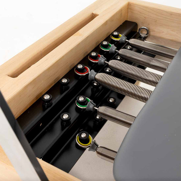 Close-up of metal springs and colored washers on black bars inside a wooden frame of the SMAI Pilates - Classical Maple Reformer, highlighting its adjustable pilates equipment.