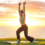 At sunset by the ocean, a person in workout gear lunges on a yoga mat, raising arms while holding SMAI Silicone Hand Weights - 1kg (Pair) with ergonomic grip design.