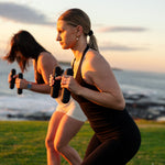 Two women exercise near the ocean at sunset, doing squats with SMAI Silicone Hand Weights - 1.5kg (Pair). Bright natural light highlights the water and grassy landscape in the background.