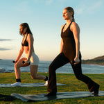 Two women exercise near the ocean at sunset, performing lunges with SMAI Silicone Hand Weights - 1kg (Pair). One wears black; the other a light sports bra and shorts. Both appear focused and active.
