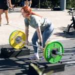 A woman with red hair is getting ready to lift a barbell equipped with SMAI's yellow and green Competition Bumper Plate 10kg (PAIR) IWF Colour outdoors on a sunny day. Dressed in a green shirt and blue leggings, she stands on raised blocks next to a stationary bike. Other people are visible in the background of this lively CrossFit scene.