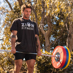 A man is outdoors engaging in weightlifting, using a barbell loaded with SMAI Competition Bumper Plates, which include IWF color pairs of red, blue, yellow, and green. Wearing a black "ZION FITNESS" t-shirt and shorts, he trains intensely while trees provide a scenic backdrop.
