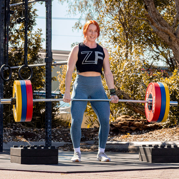 A person with red hair proudly performs a deadlift outdoors using SMAI's Competition Bumper Plate 15kg (PAIR) IWF Colour. Wearing a black crop top and blue leggings, they smile amongst the trees and exercise equipment, embodying the essence of weightlifting competitions.