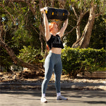 On a sunny day, a person with long red hair demonstrates their strength by lifting an SMAI Core Bag overhead in dynamic movements on an outdoor paved surface. Dressed in a black tank top and gray leggings, they are surrounded by lush greenery and trees, highlighting the rip-proof vinyl durability of the bag.