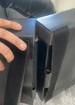 A person's hand grips two pieces of black cushioned material with Velcro, meant to be secured together, and the slightly frayed edges expose padding inside. In the background, there is a glimpse of gray flooring from the SMAI 5m Boxing Ring - Floor Mount Folding, providing context to the scene.