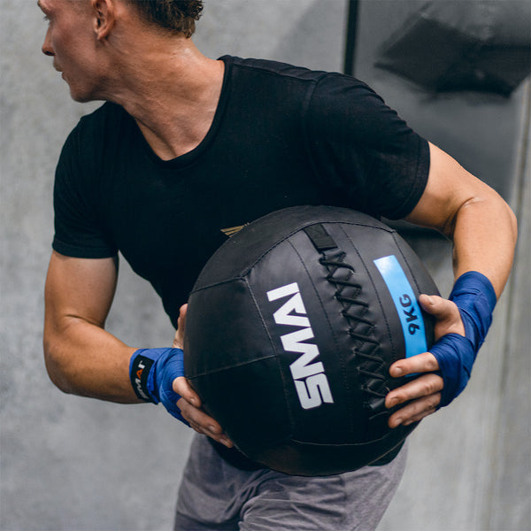 At the gym, a person in a black T-shirt and blue wrist wraps holds an SMAI Wall Ball marked "9KG" during their workout.