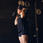 Wearing athletic clothing, a person lifts two SMAI Rubber Hex Dumbbells Classic (Pair) with precision-knurled handles at shoulder height in a gym featuring gymnastic rings and black walls.