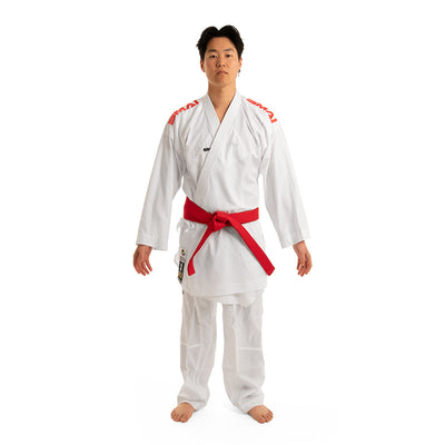 A person stands on a white background wearing the WKF Reversible Karate Uniform by SMAI, specifically the 6oz Premium Kumite Gi known as Inazuma RX, paired with a red belt. The gi features striking red logos on the shoulders that highlight focus and discipline. With their arms relaxed at their sides, they embody the true spirit of martial arts mastery.