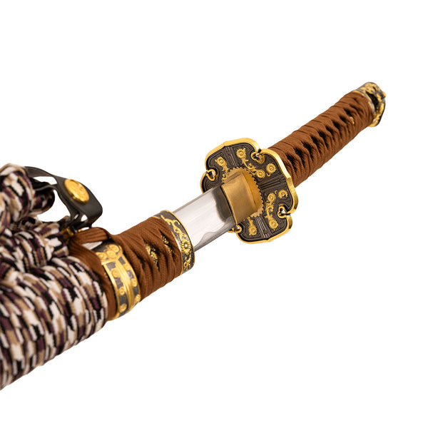 A close-up of the SMAI Forged Carbon Orange Katana - Sun Emperor handle, featuring intricate gold and black details, brown wrapping, a patterned cord, and an ornate tsuba (sword guard) design.