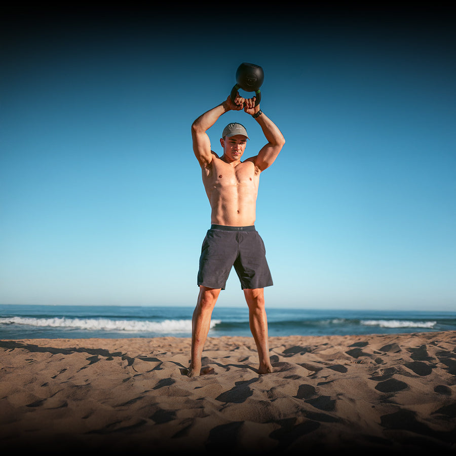 A shirtless man wearing a cap and shorts stands barefoot on a sandy beach, lifting a kettlebell overhead. The sky is clear and ocean waves are visible in the background.