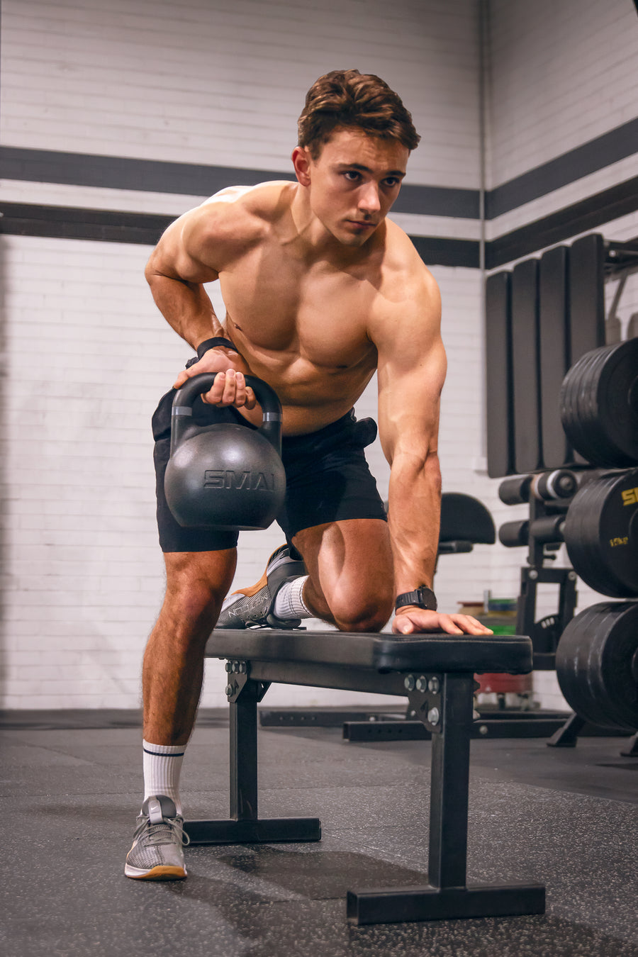 A shirtless man does a single-arm row with a kettlebell while kneeling on a bench in a gym, surrounded by weightlifting equipment.