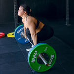 An athlete in a gym deadlifts with a barbell fitted with SMAI 20kg Competition Bumper Plates in IWF colors. Focused and poised, they grip the weights, knees bent and back straight, ready to lift the barbell like in weightlifting competitions.