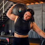 Wearing athletic gear, a person lifts an 80kg SMAI Slam Ball Monster onto their shoulder in a gym, showcasing focus and effort. Gym equipment and stairs appear in the background.