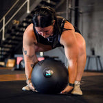 Wearing athletic gear, a person crouches to lift the SMAI Slam Ball Monster - 80kg in a gym with stairs and equipment visible in the background.