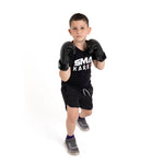 A young boy wearing SMAI Essentials Kids Boxing Glove - Black, a black "SMA Karate" t-shirt, black shorts, and sneakers stands in a fighting stance against a plain white background.