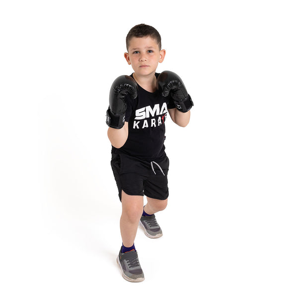 A young boy wearing SMAI Essentials Kids Boxing Glove - Black, a black "SMA Karate" t-shirt, black shorts, and sneakers stands in a fighting stance against a plain white background.