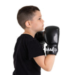 A young boy in a black t-shirt wears SMAI Essentials Kids Boxing Glove - Black, standing in a boxing stance with hands raised near his face, facing sideways against a plain white background.