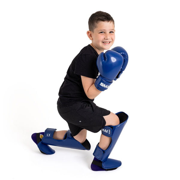 A smiling young boy kneels on one knee, wearing a black t-shirt and shorts with SMAI Essentials Kids Boxing Glove - Blue, posing against a plain white background.