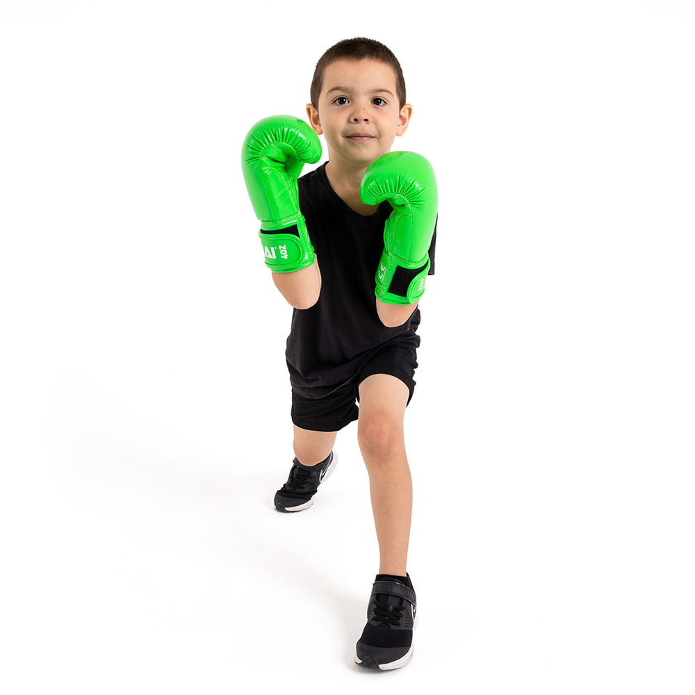 A young boy in a black t-shirt and shorts stands in a boxing stance with one foot forward, wearing bright green SMAI Essentials Kids Boxing Gloves, set against a plain white background.