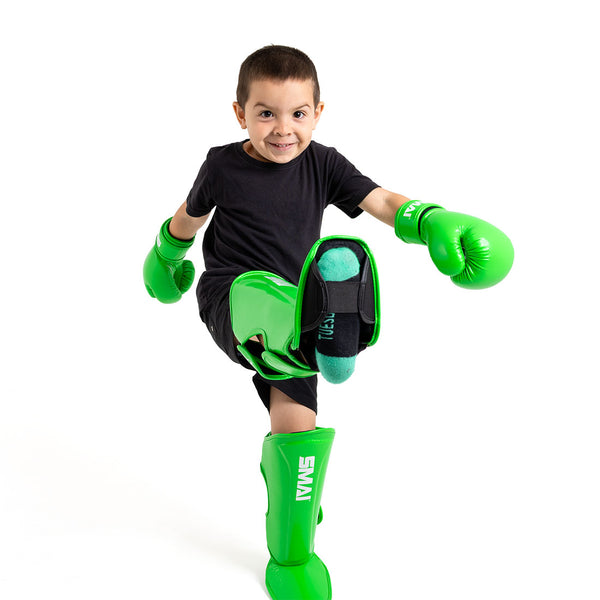 A young boy in black clothing and bright green SMAI Essentials Muay Thai Shin Guards for kids smiles energetically and kicks forward, wearing boxing gloves against a plain white background.