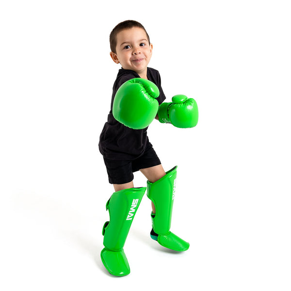 A child in black poses playfully in bright green SMAI Essentials Muay Thai Shin Guards for kids, ready in a boxing stance against a white background, highlighting protective children's gear.
