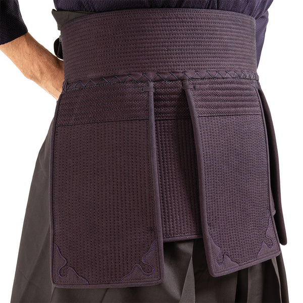 A close-up shows a person wearing the dark indigo tare waist and hip padding from the SMAI Kendo Bogu Set, part of traditional Japanese martial arts armor, over pleated trousers.