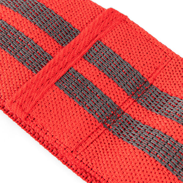 A close-up shot features a red knitted mini resistance band from the No Sync To Channel Engine's set of three, adorned with two parallel black stripes. Its coarse texture reveals a woven pattern. The sharp focus on the foreground highlights the band’s intricate details, making it ideal for glute activation, while the softly blurred background enhances its prominence.