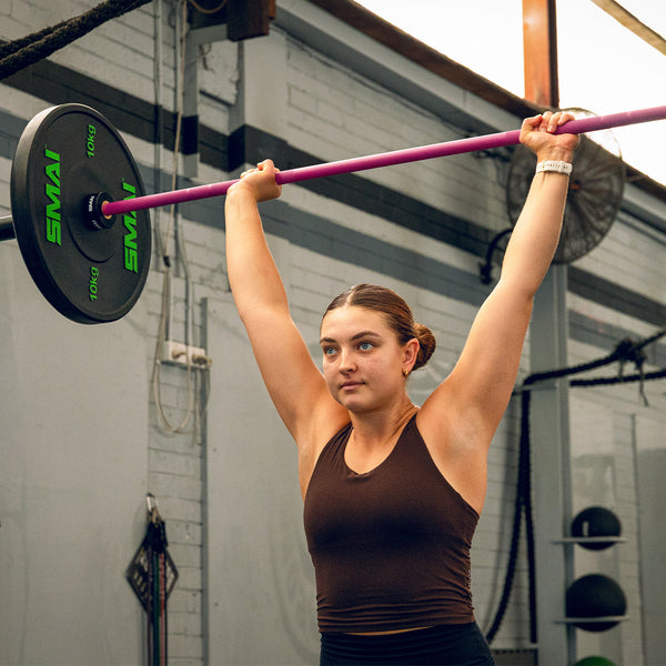 A woman in athletic wear lifts a 20kg SMAI Barbell - Magenta above her head in a gym, looking focused as she stands before gym equipment and a white brick wall.