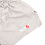 Close-up of SMAI Muay Thai Shorts - Grey, unisex satin polyester with elastic waistband and "SMAI" embroidered near the hem—the "A" in red, others white.