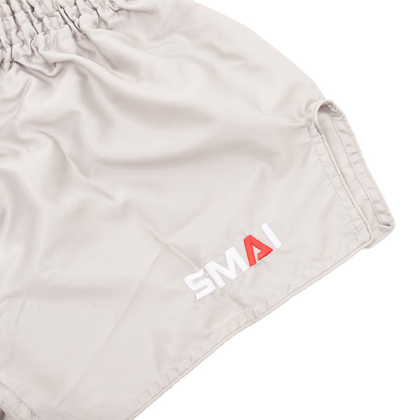 Close-up of SMAI Muay Thai Shorts - Grey, unisex satin polyester with elastic waistband and "SMAI" embroidered near the hem—the "A" in red, others white.