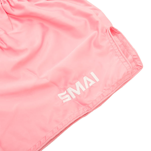 SMAI Muay Thai Shorts - Pink: Light pink satin polyester with "SMAI" white embroidery on the lower leg, shown in a close-up highlighting the fabric and stitching details.