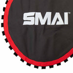 Close-up of a round Ubound Trampoline - Mat from No Sync To Channel Engine, featuring a dark surface with red trim and bold white "SMAI" lettering. This model includes spare parts like a replacement mat for maintaining optimal performance, with black webbing surrounding the trampoline edge.