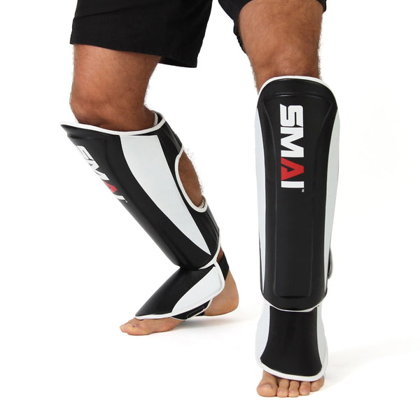 A person stands barefoot against a white background, wearing SMAI Essentials Muay Thai Shin Guards in black and white (XL), made from synthetic leather with EVA foam protection.