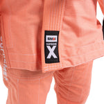 Close-up of the SMAI Supreme X Brazilian Jiu Jitsu Uniform in salmon, featuring a "SMAI SUPREME X" jacket patch and "SUPREME" vertically on the left pant leg—perfect for youth martial arts.