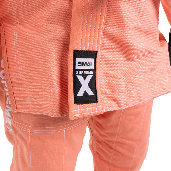 Close-up of the SMAI Supreme X Brazilian Jiu Jitsu Uniform in salmon, featuring a "SMAI SUPREME X" jacket patch and "SUPREME" vertically on the left pant leg—perfect for youth martial arts.