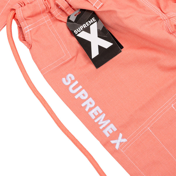 Close-up of the Supreme X Brazilian Jiu Jitsu Uniform - Salmon by SMAI. The coral pink drawstring pants feature white stitching, "SUPREMEX" embroidery, and a black "SUPREME X" tag on the waistband. Youth martial arts-inspired style.
