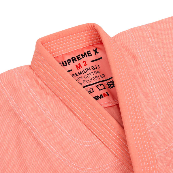 Close-up of the SMAI Supreme X Brazilian Jiu Jitsu Uniform - Salmon, a pink pearl weave youth Gi jacket with white stitching. Inner label shows size M2, 65% cotton/35% polyester blend, plus care instructions.