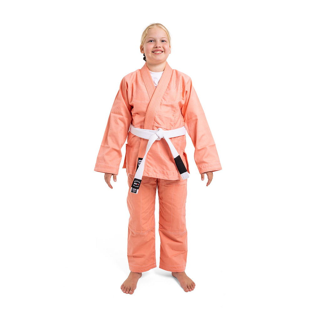 A smiling child wears the SMAI Supreme X Brazilian Jiu Jitsu Uniform - Salmon with a white belt, standing barefoot against a plain white background.