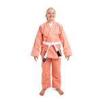 A smiling child wears the SMAI Supreme X Brazilian Jiu Jitsu Uniform - Salmon with a white belt, standing barefoot against a plain white background.