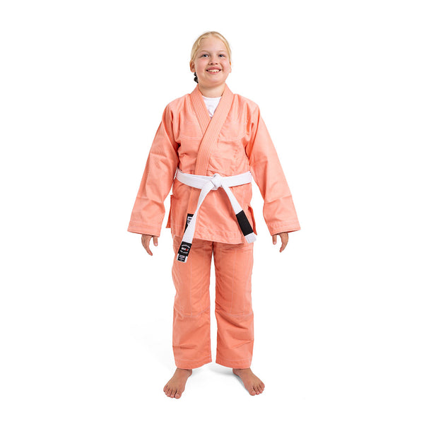 A smiling child wears the SMAI Supreme X Brazilian Jiu Jitsu Uniform - Salmon with a white belt, standing barefoot against a plain white background.