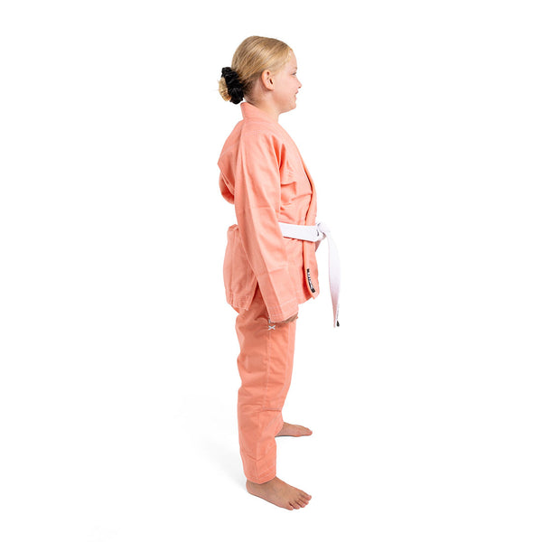 A young girl stands barefoot in side profile, wearing the SMAI Supreme X Brazilian Jiu Jitsu Uniform in Salmon with a white belt, crafted from pearl weave fabric, against a plain white background.