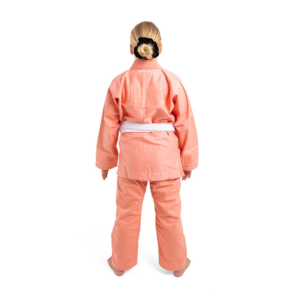 A person with blonde hair in a bun, wearing the SMAI Supreme X Brazilian Jiu Jitsu Uniform in salmon and a white belt, stands barefoot facing away against a white background.