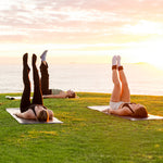 Three people work out on SMAI Yoga Mat / Pilates Mat - Dark Grey on the grass by the ocean at sunset, raising their legs as part of their exercise routine with an orange sky and low sun in the background.