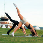 Two women practice yoga outdoors on grass in a downward dog split pose. One uses the SMAI Yoga Mat / Pilates Mat - Dark Grey, known for its non-slip grip, while the other wears beige sports bra and shorts; buildings and tents are visible behind them.